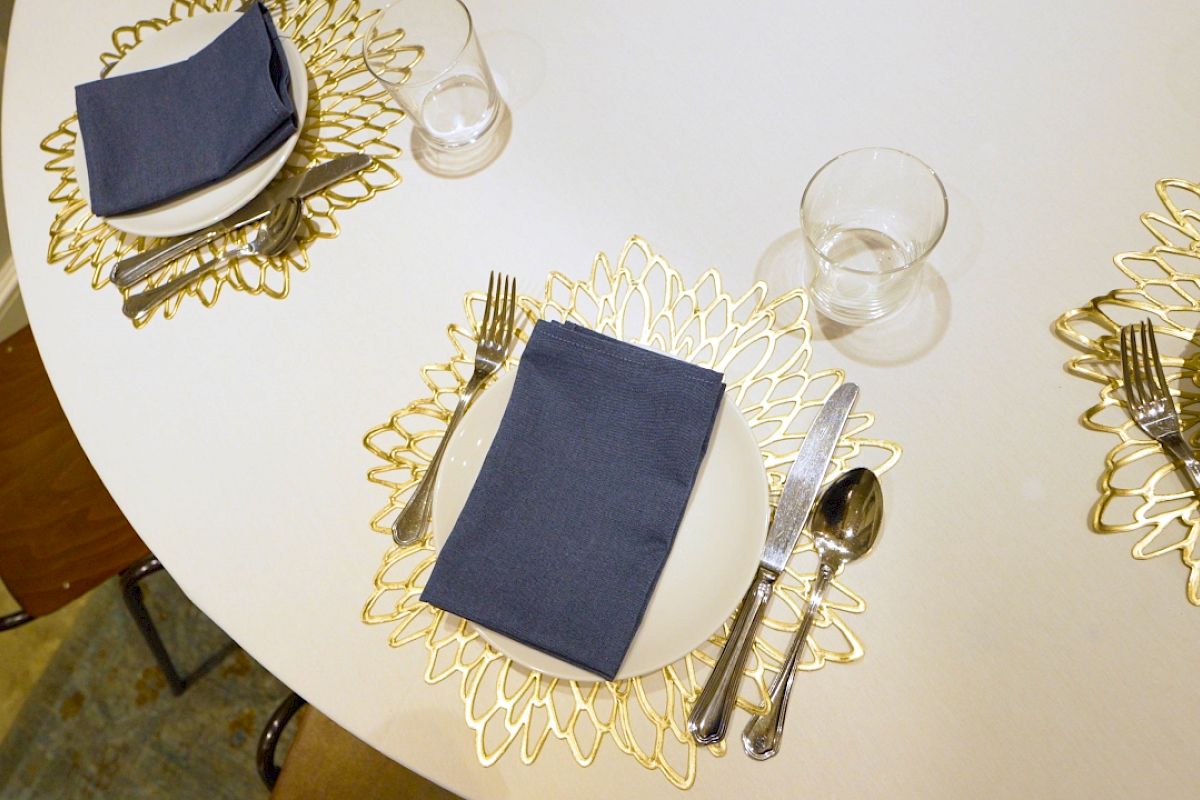 A round table set with plates, cutlery, glasses, and neatly folded blue napkins on decorative gold placemats, prepared for a meal.