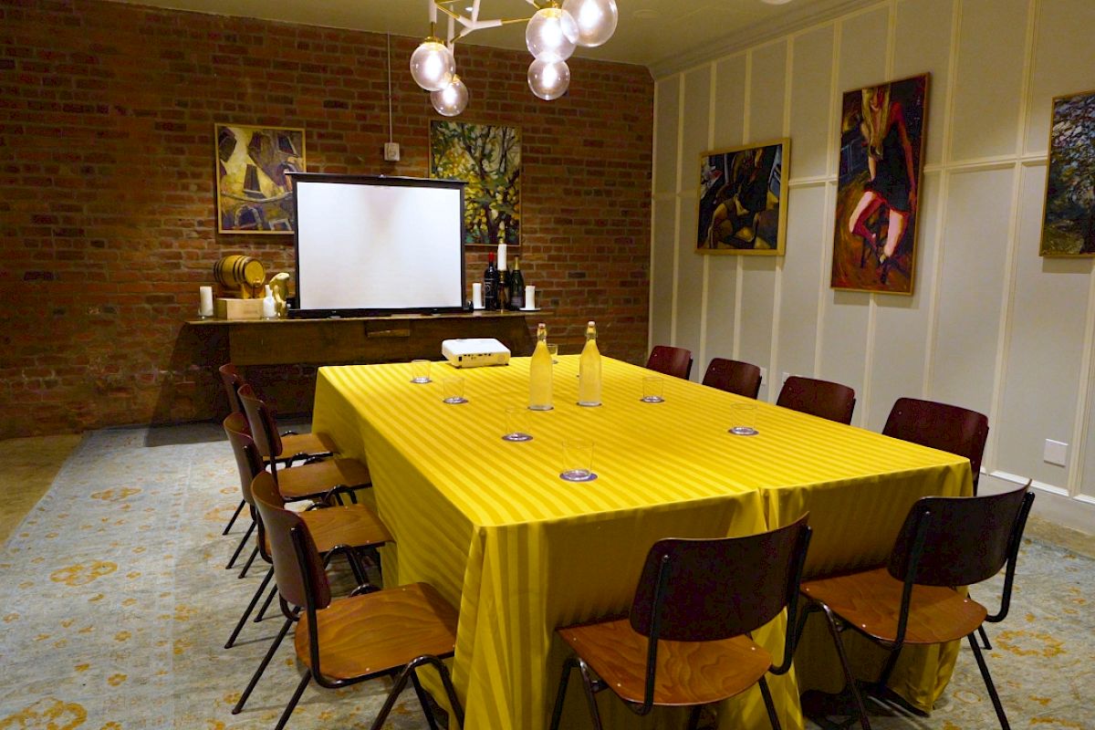 A meeting room features a yellow tablecloth, chairs, a projector, and artworks on brick and white panel walls, with bottles and coasters on the table.