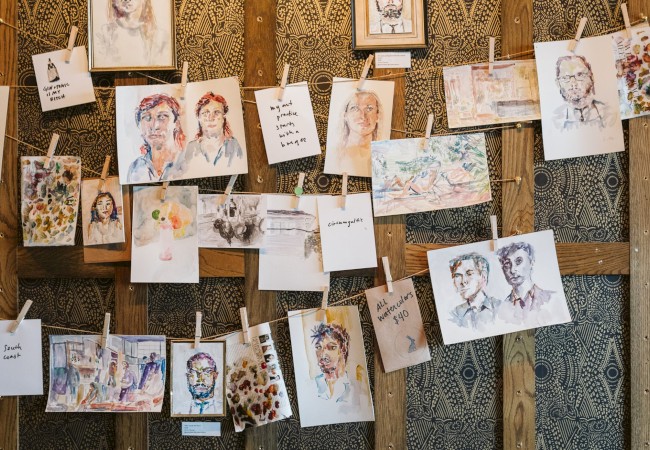 The image shows a display of various paintings and sketches clipped to strings on a wooden wall.
