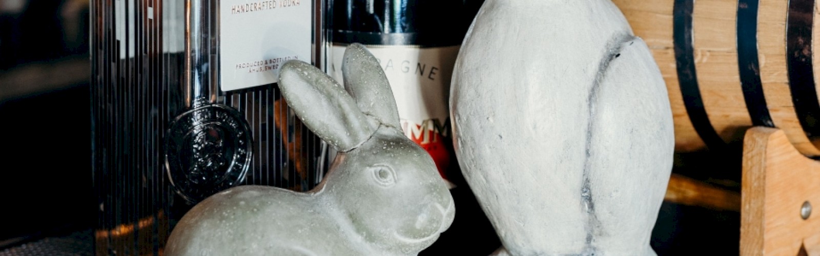 The image shows two rabbit statues, a bottle of gin, a bottle of Champagne, and a small wooden barrel on a table.