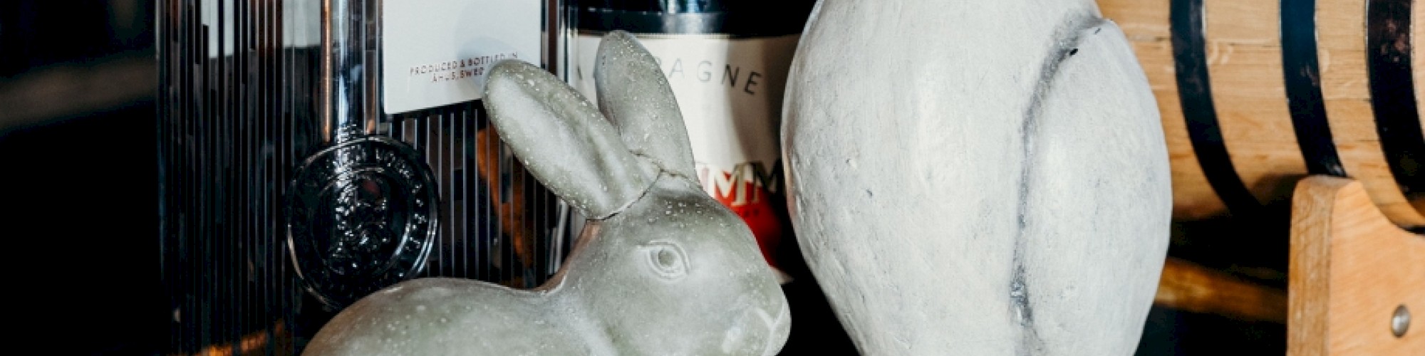 The image shows two rabbit statues, a bottle of gin, a bottle of Champagne, and a small wooden barrel on a table.