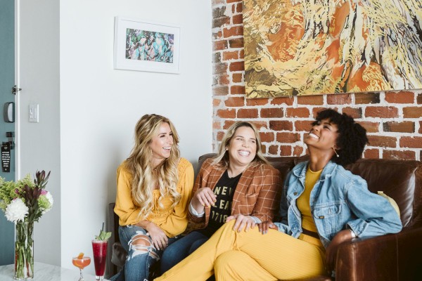 Three people sit on a couch in a cozy room with artwork on the brick wall, a table with flowers and drinks in front of them, and they appear to be smiling.