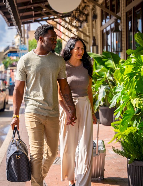 A stylish couple walks hand-in-hand down a sunny outdoor shopping street, stylish in light beige outfits, smiling and chatting.
