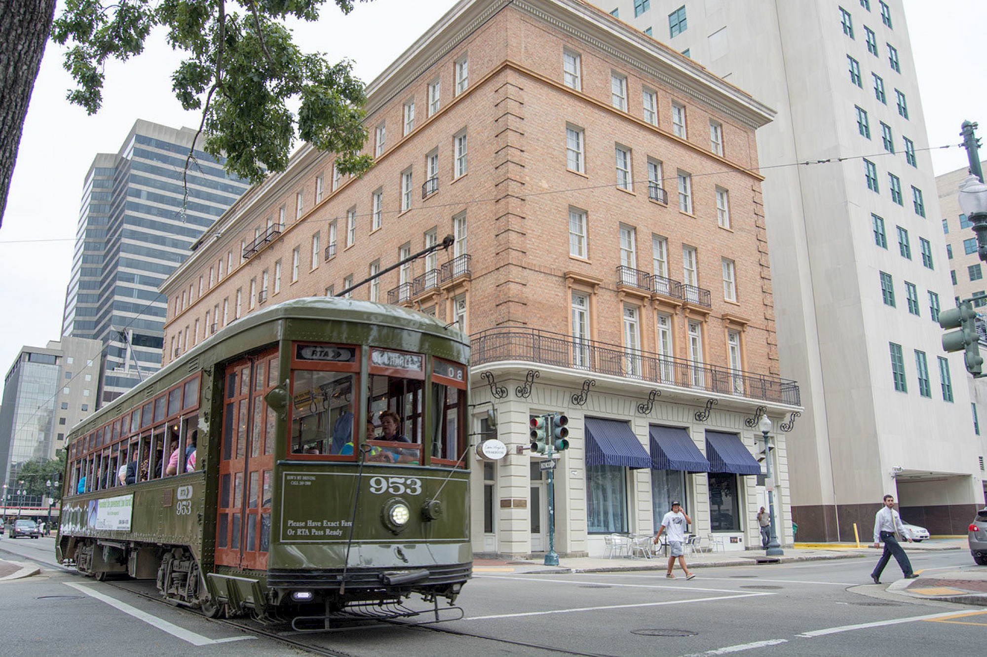 A green streetcar numbered 953 is seen traveling along a city street with pedestrians walking nearby and buildings in the background.