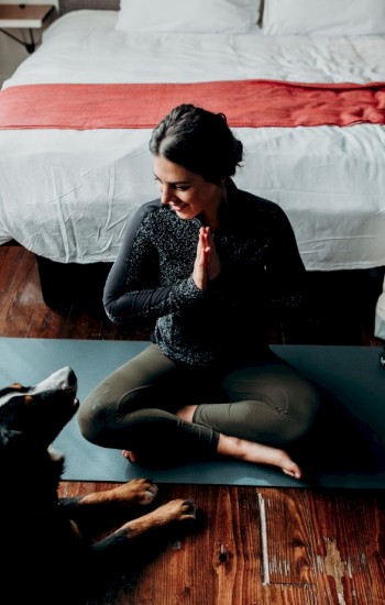 A woman sits in a yoga pose on a mat with a dog in front of her. A bed and tote bag are in the background, ending the sentence.