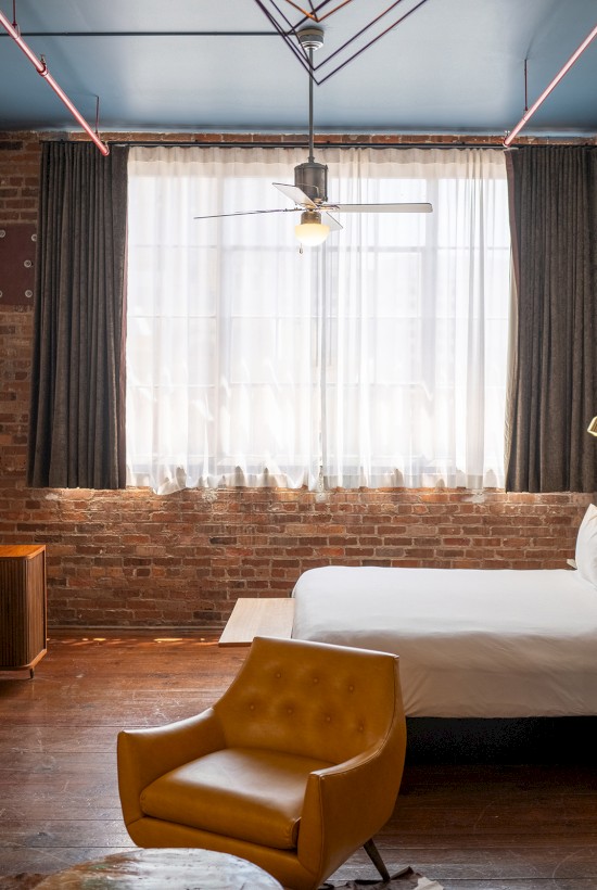The image shows a stylish hotel room with a large bed, armchair, and wooden furniture, featuring brick walls and industrial decor elements.