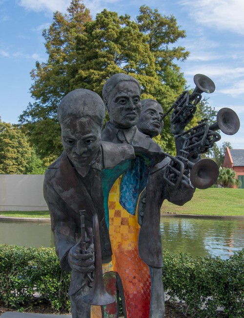 Statue of three musicians playing brass instruments beside a pond with a bridge and greenery, and a building in the background on a sunny day.