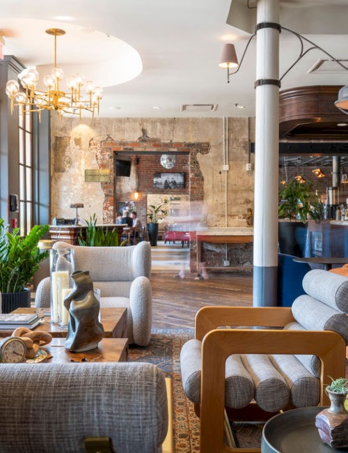 A stylish, modern lounge with eclectic furniture, large windows, and a bar in the background. Indoor plants and decorative pieces are present.