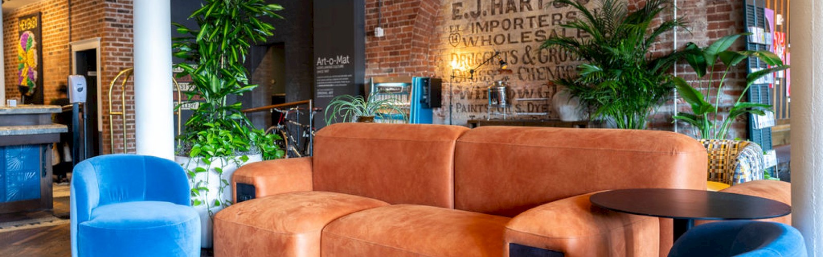The image shows a cozy lounge area with an orange sofa, blue chairs, wooden flooring, plants, and a brick wall with vintage signage in the background.