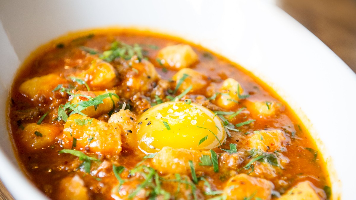 A bowl of stew with chunks of meat, vegetables, herbs, and a whole egg on top, garnished with green herbs on a wooden table.