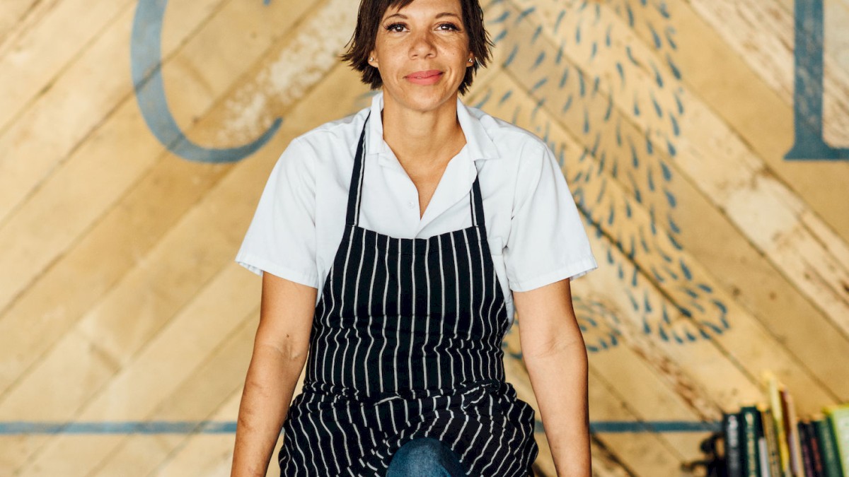 A person wearing an apron sits on a bar counter, leaning back. The background features a rustic wall design with a rabbit illustration and partial text.