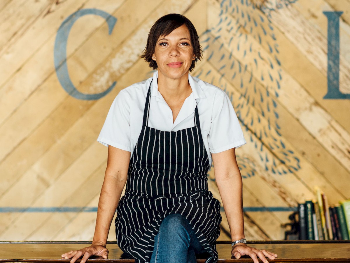 A person wearing an apron sits on a bar counter, leaning back. The background features a rustic wall design with a rabbit illustration and partial text.