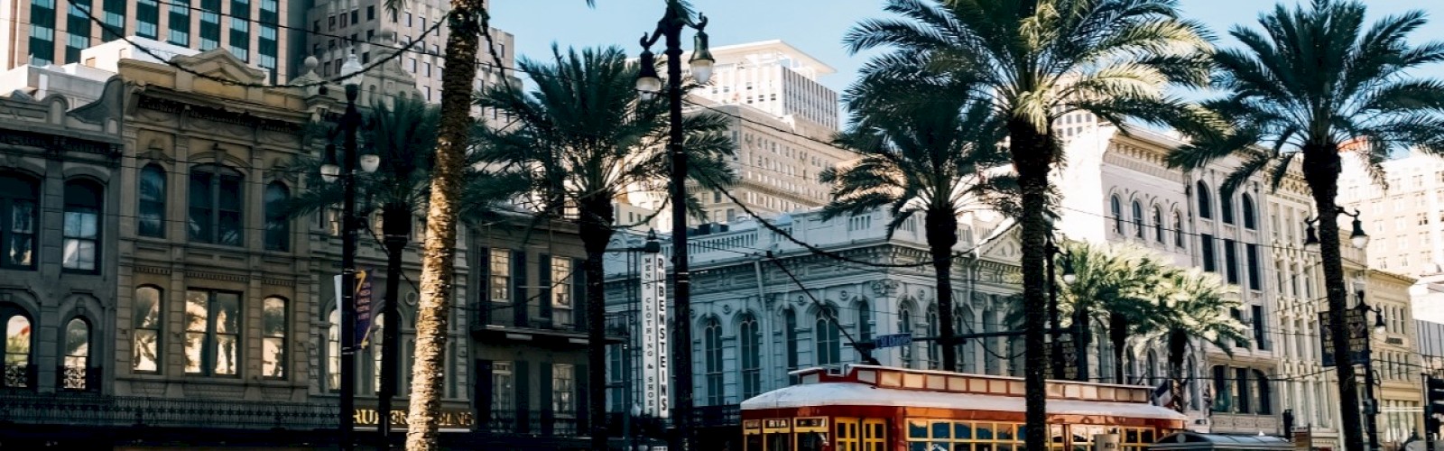The image shows a street with palm trees, historic buildings, and a red streetcar under a clear blue sky, capturing an urban atmosphere.