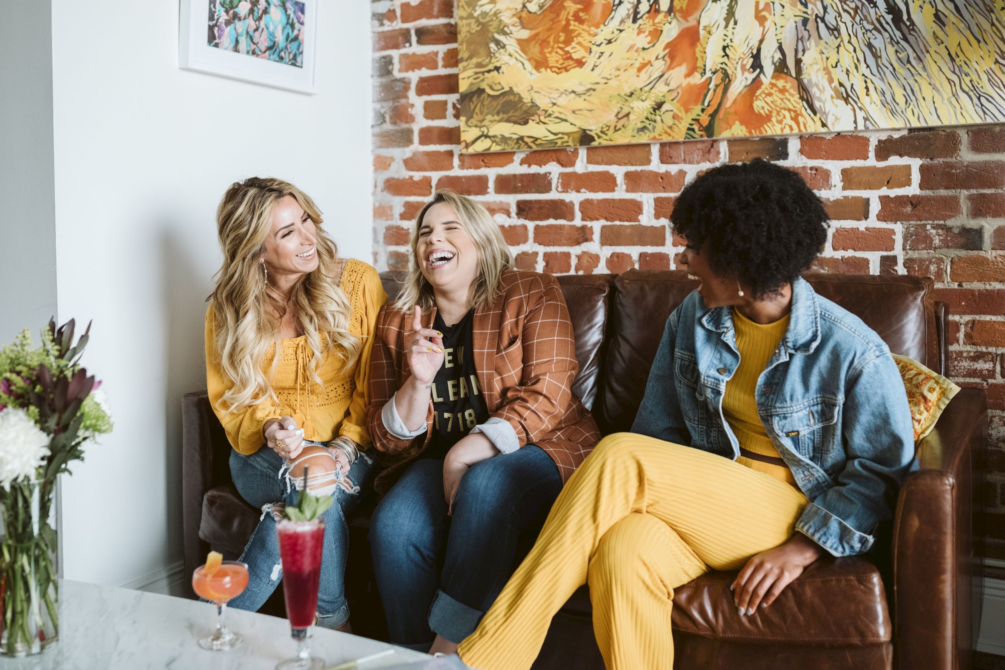 Three women sitting on a couch laughing and talking, with drinks on the table and artwork in the background.