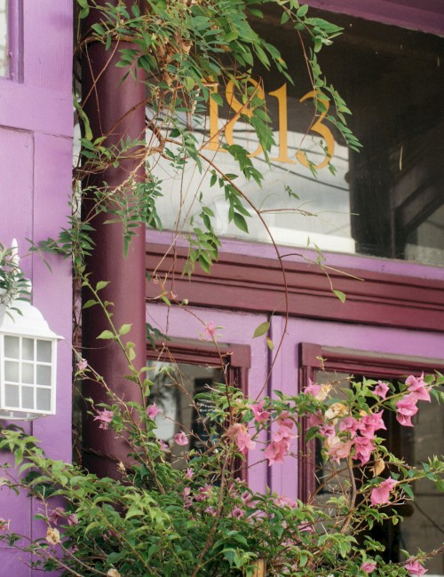 A purple building facade with the number 812, a lantern-style light fixture, and pink flowering plants climbing up the wall.