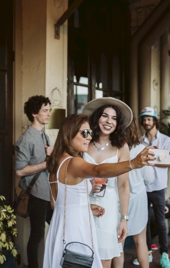 A group of people, mostly in white outfits, are gathered outside a building. Two women take a selfie, and others are casually chatting.