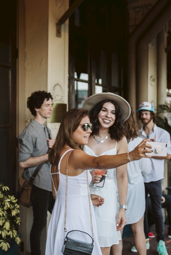 A group of people, mostly in white outfits, are gathered outside a building. Two women take a selfie, and others are casually chatting.