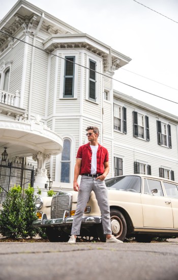 A man in a red shirt and jeans stands in front of a vintage car with luggage, beside a large, white Victorian-style house.