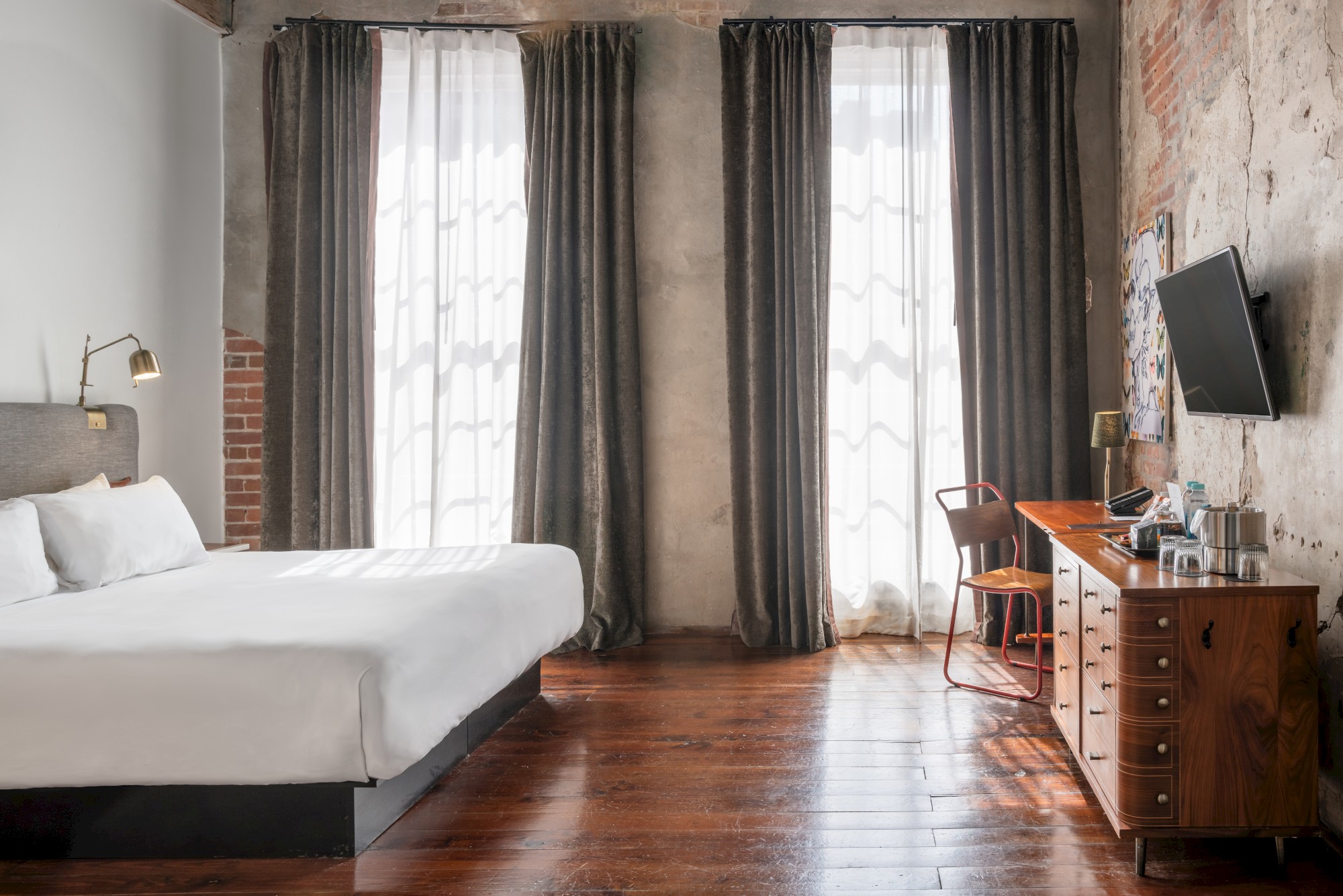 A modern hotel room with a bed, desk, chair, TV, and windows with dark curtains. The floor is wooden and the walls have an industrial style.