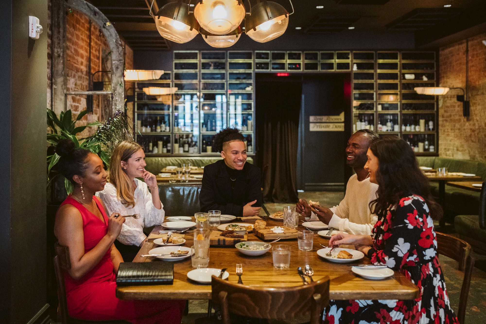 Five people are sitting around a table in a cozy restaurant, enjoying food and engaging in conversation, with shelves and warm lighting in the background.