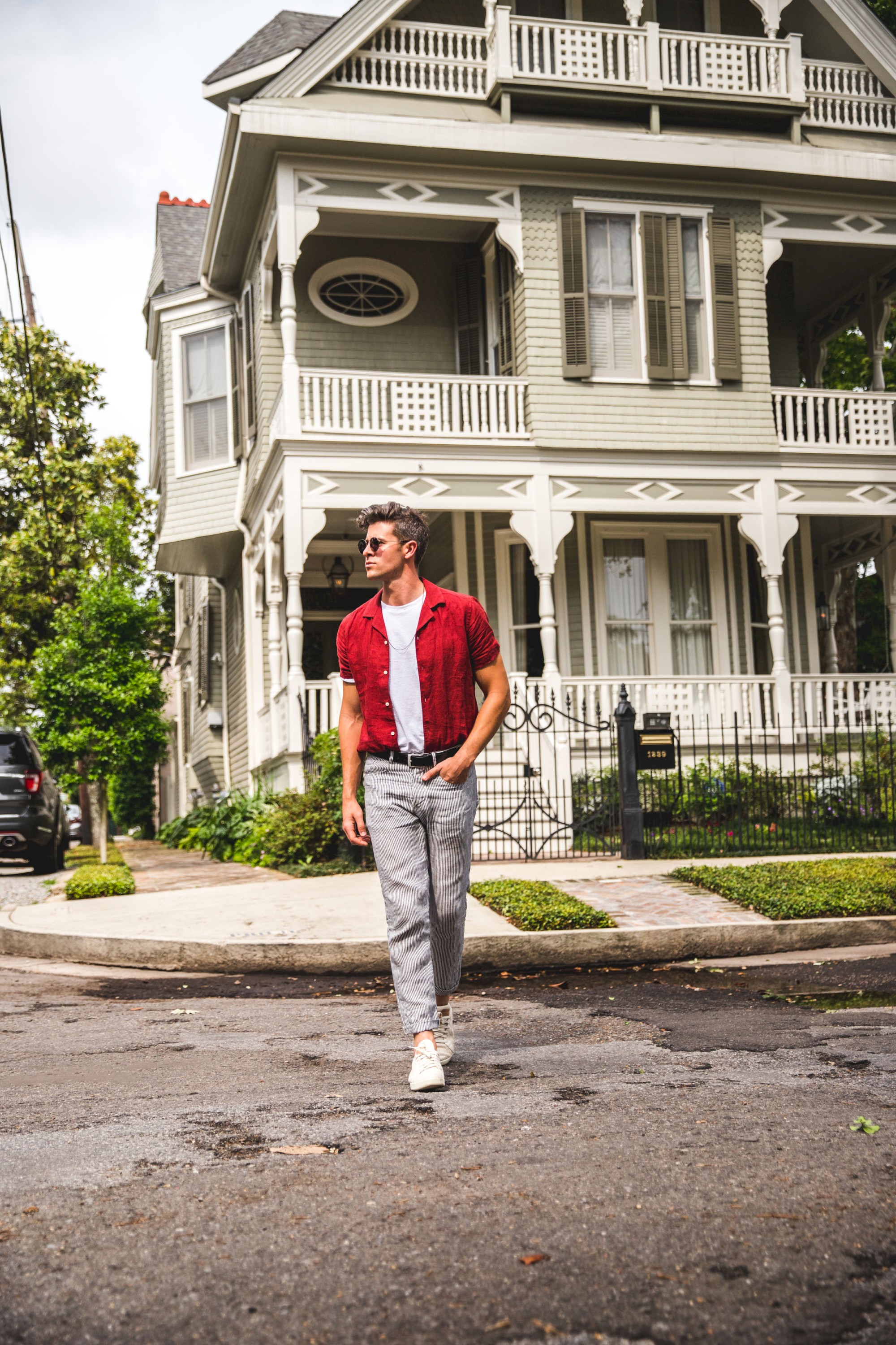 A person walks on a street in front of a large, ornate house with a porch and bay windows, wearing a red shirt and sunglasses.