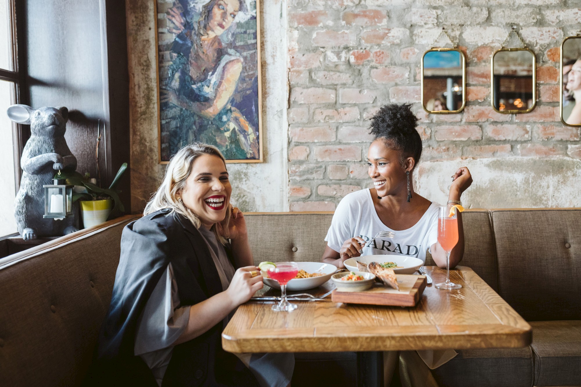 Two people are enjoying drinks and snacks at a cozy cafe, sitting at a wooden table with art on the brick wall and mirrors.