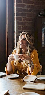 A person is sitting at a wooden table in a restaurant, holding a cup and smiling, with a brick wall backdrop and framed artwork.