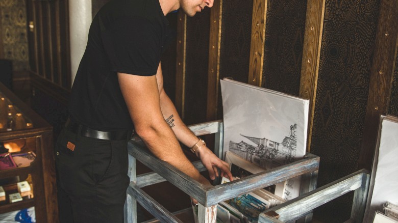 A man is examining artwork in a gallery, with paintings displayed on the wall and prints in bins.