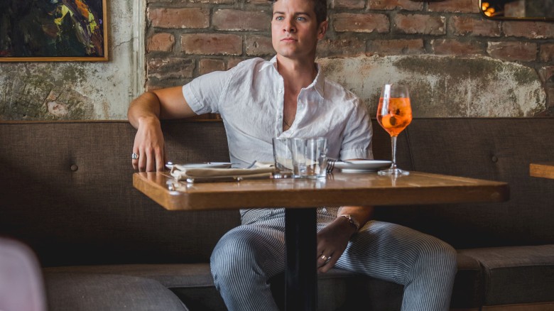 A person sitting at a cozy restaurant table with a drink, brick wall, and artwork in the background.