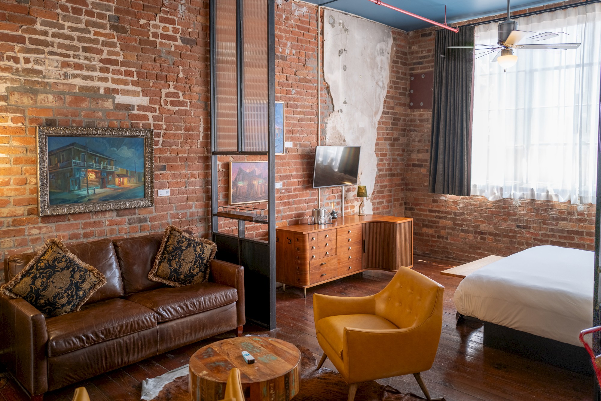 A cozy room with exposed brick walls, stylish furniture, a leather sofa, a yellow chair, and a bed. It has a modern rustic ambiance.