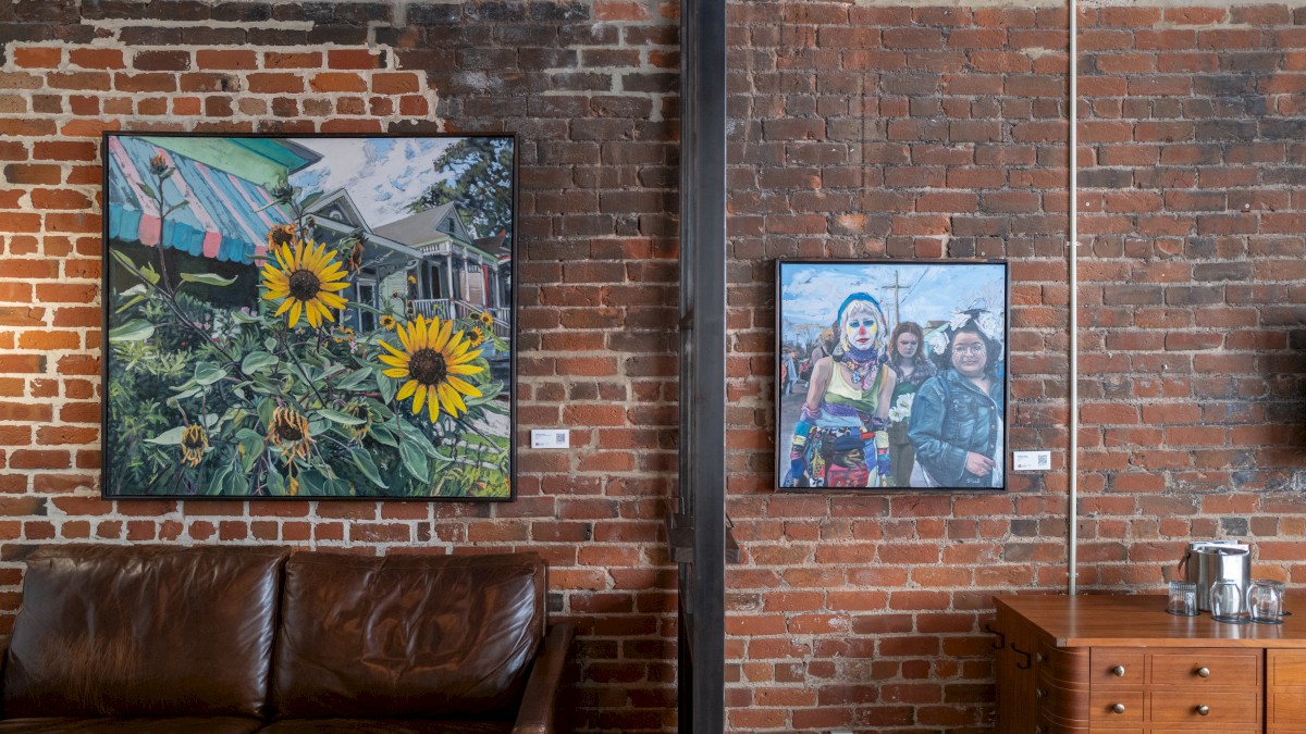 The image shows a brick wall with two paintings: one of sunflowers, the other of people. Below are a brown sofa and a wooden drawer.