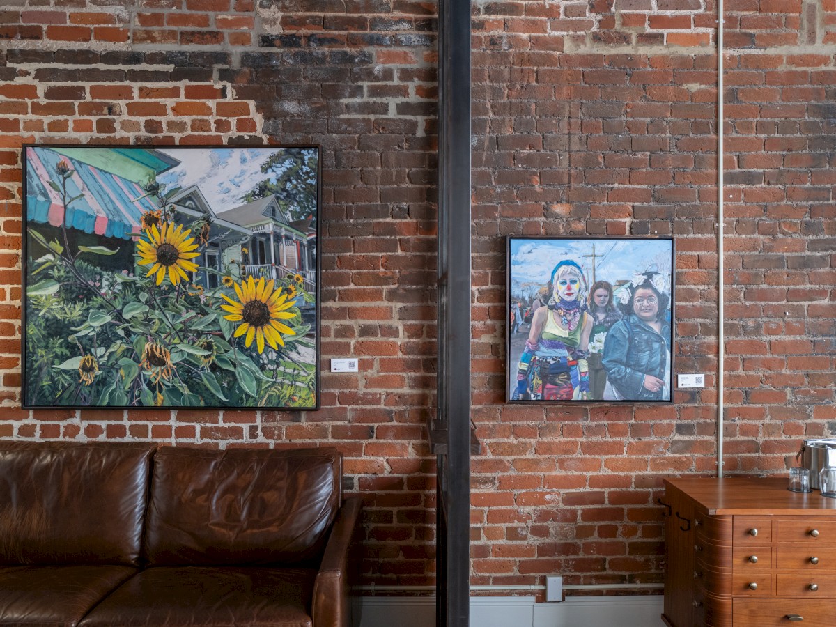 The image shows a brick wall with two paintings: one of sunflowers, the other of people. Below are a brown sofa and a wooden drawer.