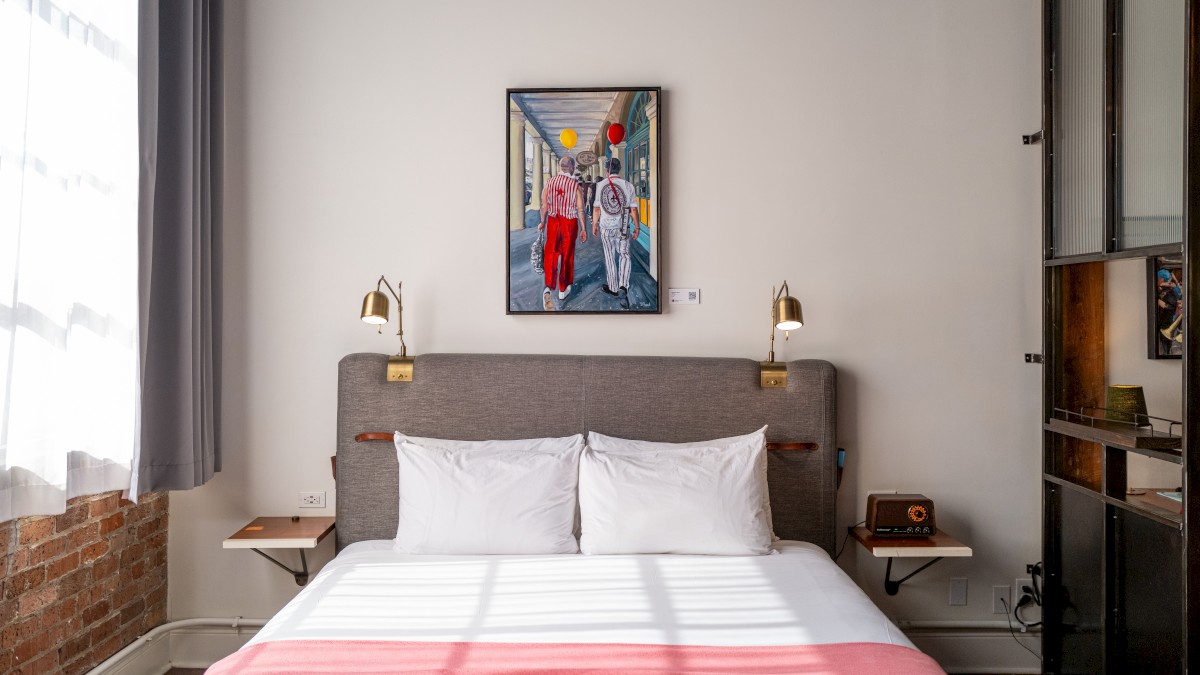 A cozy bedroom features a neatly made bed, wall lamps, a vintage phone, and colorful artwork above the headboard, with sunlight streaming in.