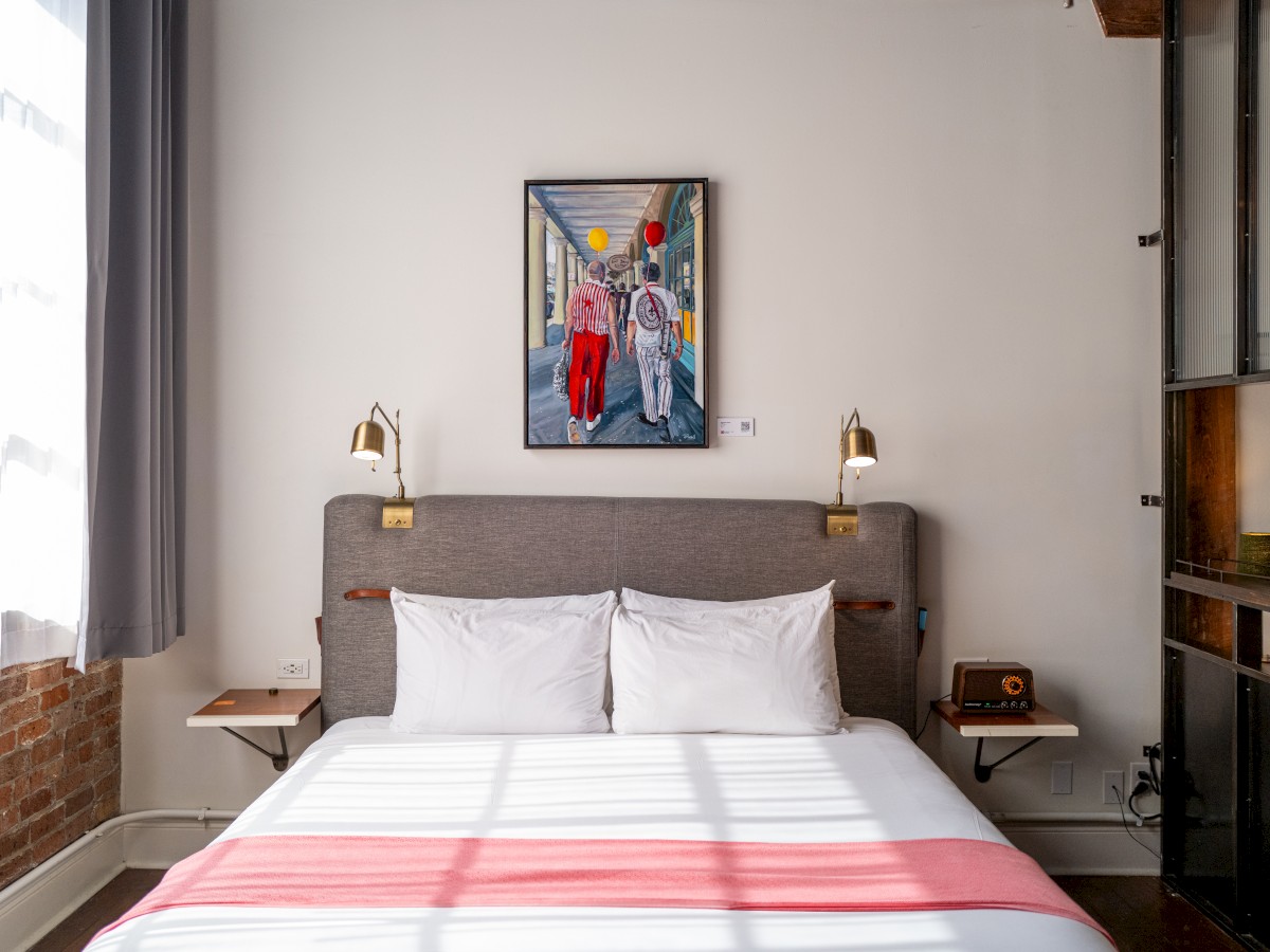 A cozy bedroom features a neatly made bed, wall lamps, a vintage phone, and colorful artwork above the headboard, with sunlight streaming in.