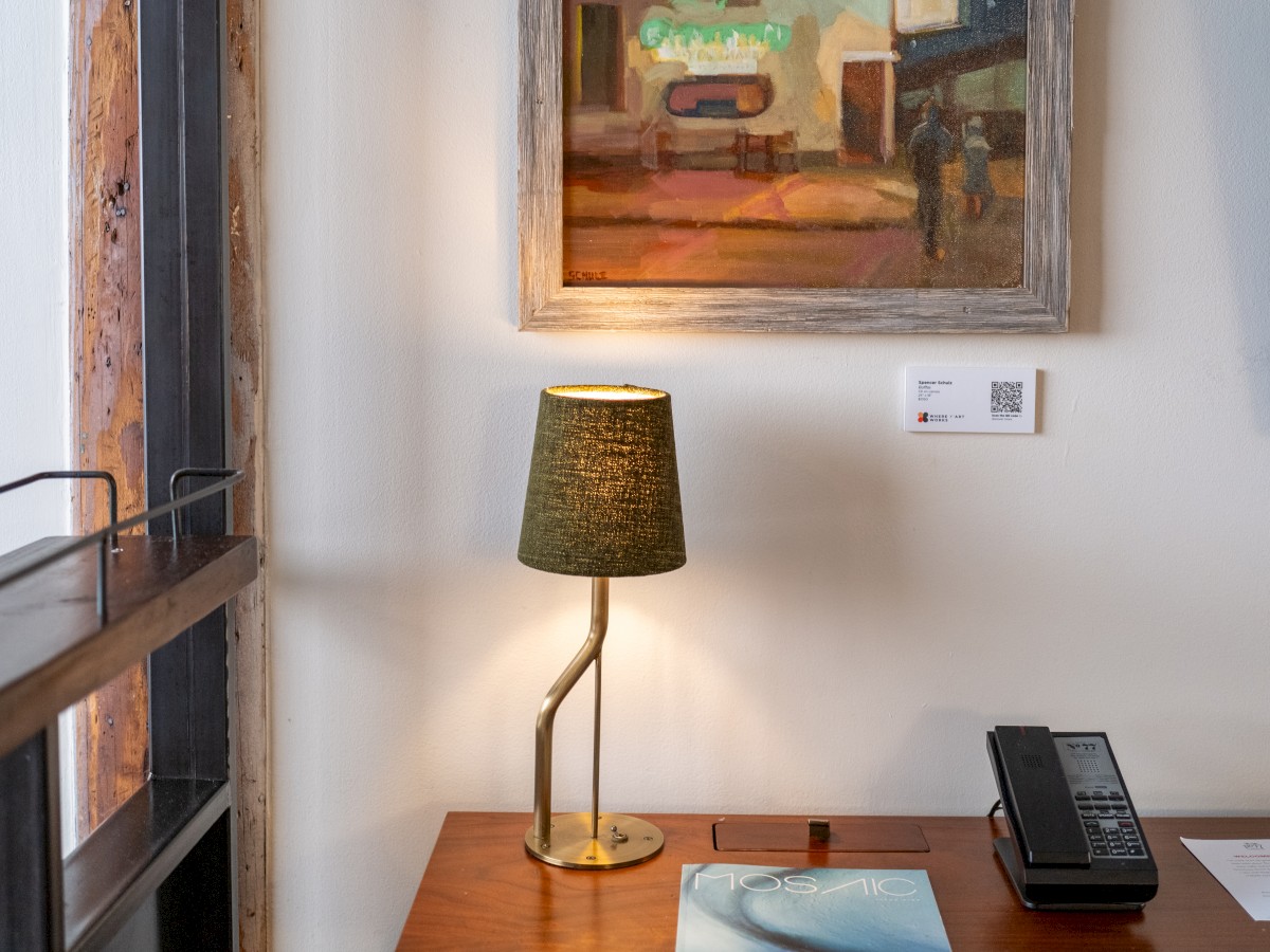 A cozy workspace featuring a wooden desk with a lamp, phone, and magazine. Above is a framed painting. A red chair is in front of the desk.