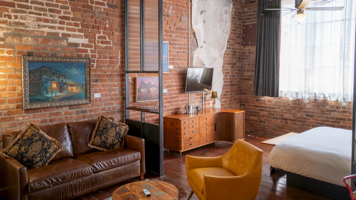 A cozy room with brick walls features a leather sofa, yellow chair, bed, TV, and a wooden dresser, creating a stylish, rustic ambiance.