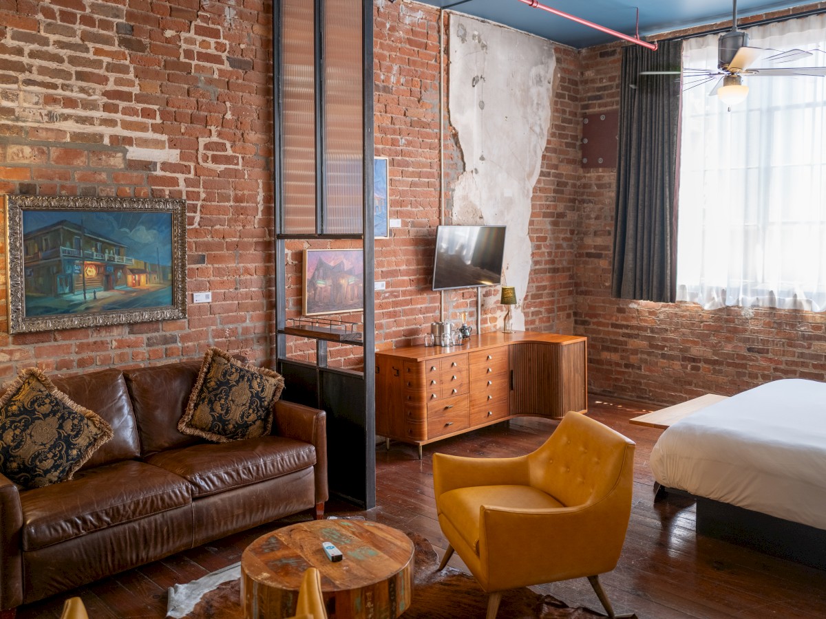 A cozy room with brick walls features a leather sofa, yellow chair, bed, TV, and a wooden dresser, creating a stylish, rustic ambiance.