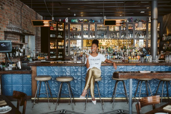 A woman sits at a stylish bar with blue paneling, surrounded by shelves of bottles, high stools, and warm lighting.