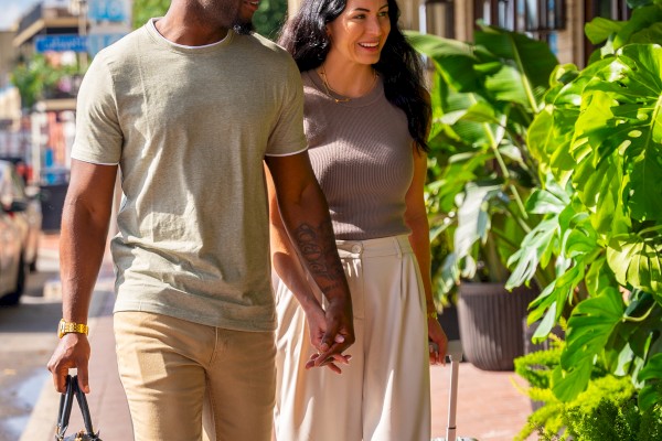 A couple walks on a sunny street, surrounded by greenery, holding luggage bags and holding hands.