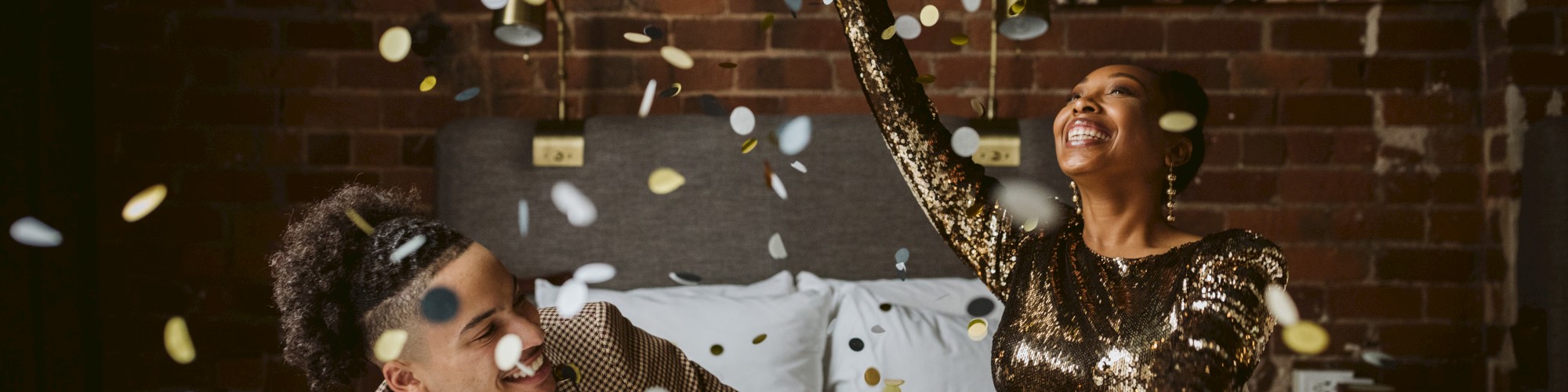 Two people joyfully toss confetti while sitting on a bed, with a brick wall and artwork in the background.