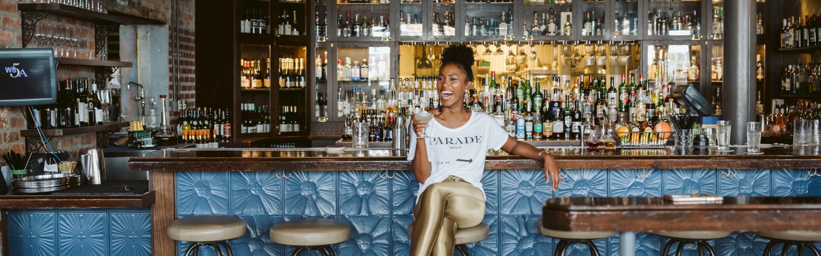 A person is sitting at a bar counter, surrounded by stools, shelves of bottles, and a warm, stylish interior.