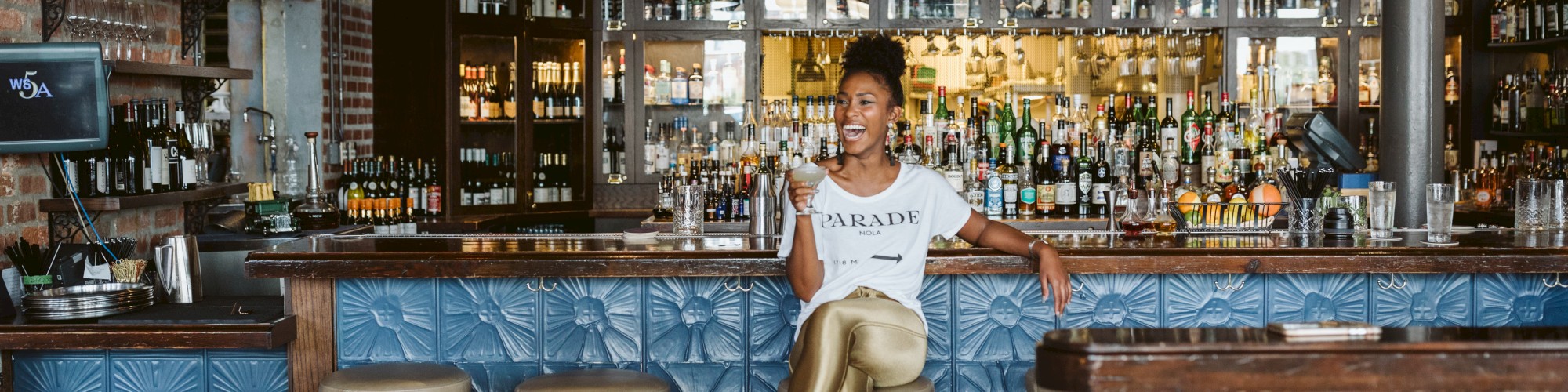 A person is sitting at a bar counter, surrounded by stools, shelves of bottles, and a warm, stylish interior.