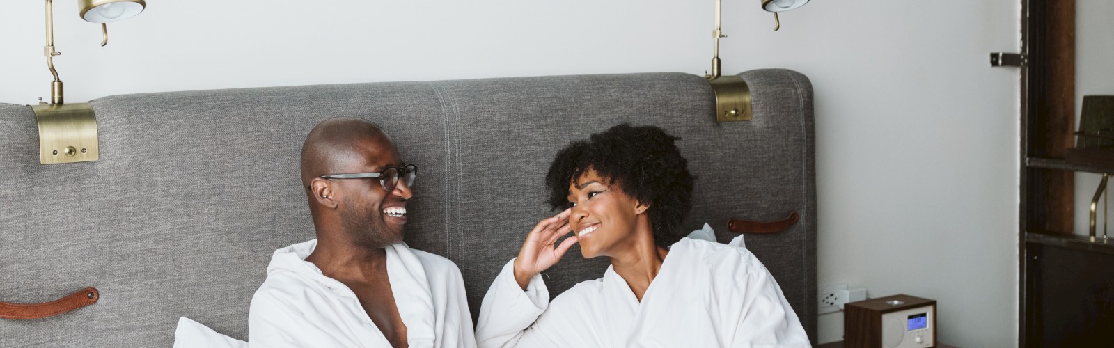 Two people in white robes are sitting on a bed in a room, smiling at each other with a blue abstract painting above them.