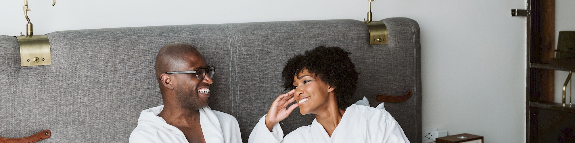 Two people in white robes are sitting on a bed in a room, smiling at each other with a blue abstract painting above them.