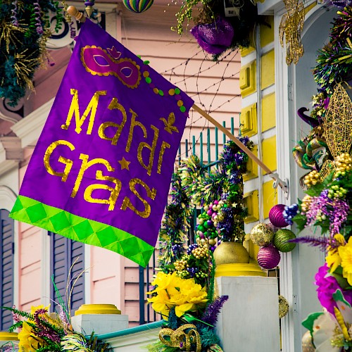 The image shows a festive Mardi Gras-themed display with a purple flag and decorations in yellow, gold, and green on a building facade.