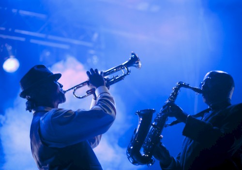 Two musicians perform on stage, one playing a trumpet and the other a saxophone, illuminated by blue lighting and smoke effects.