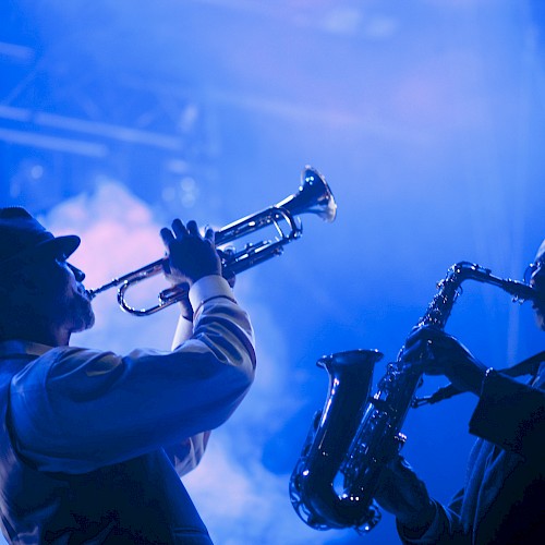 Two musicians perform on stage, one playing a trumpet and the other a saxophone, illuminated by blue lighting and smoke effects.