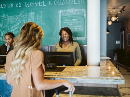 A woman with a suitcase at a hotel front desk, two receptionists assisting her in a lobby with a chalkboard wall.