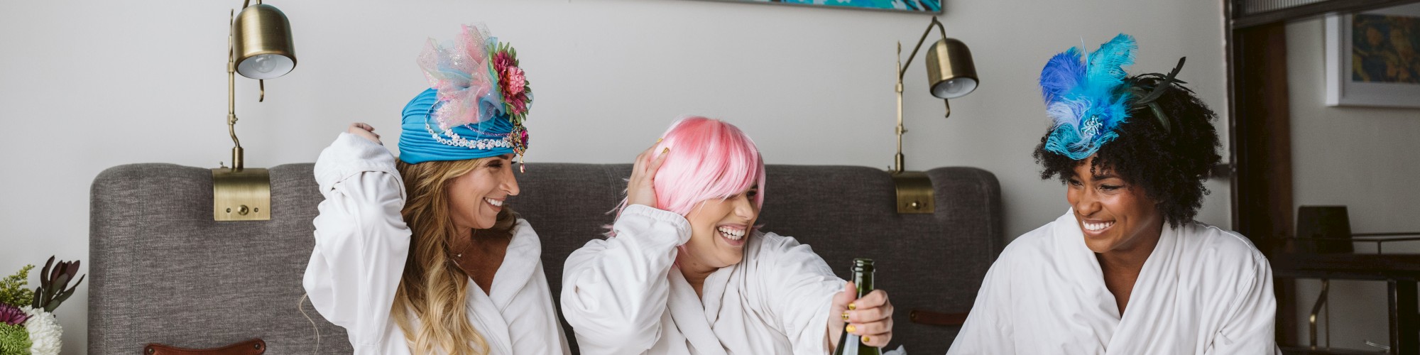 Three people in robes and colorful hats are on a bed, holding a bottle, with a vibrant painting on the wall behind them.