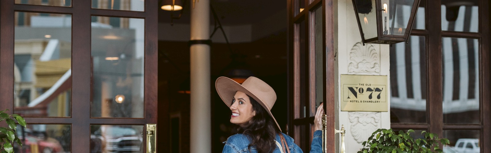 A person wearing a hat and denim jacket enters a stylish building with large glass doors and potted plants outside.